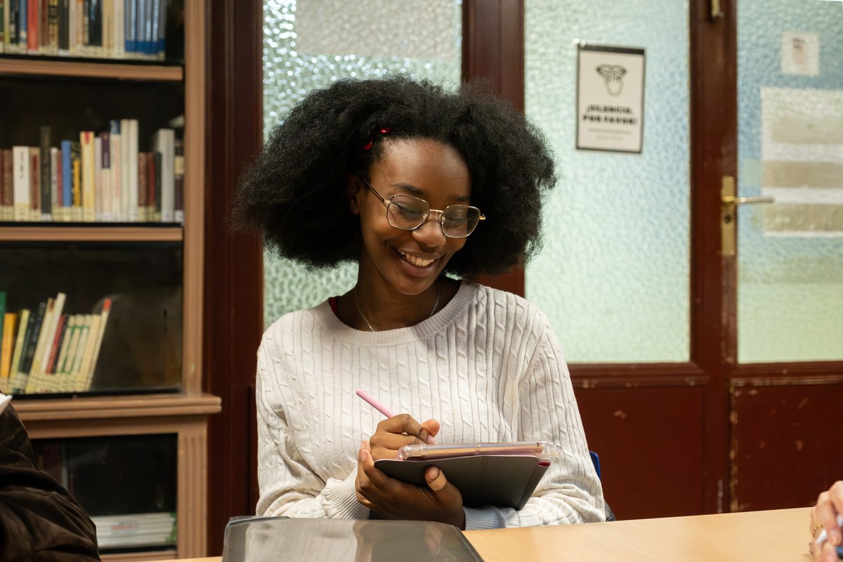Smiling young woman with glasses studying with a tablet beside bookshelves