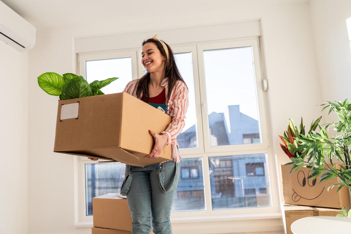 Smiling woman carrying a moving box with a plant into a bright new flat