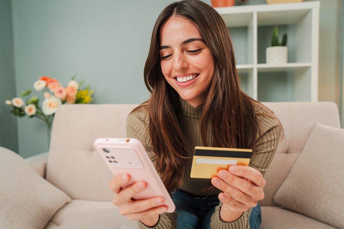 Smiling woman holding a phone and a bank card on a sofa at home
