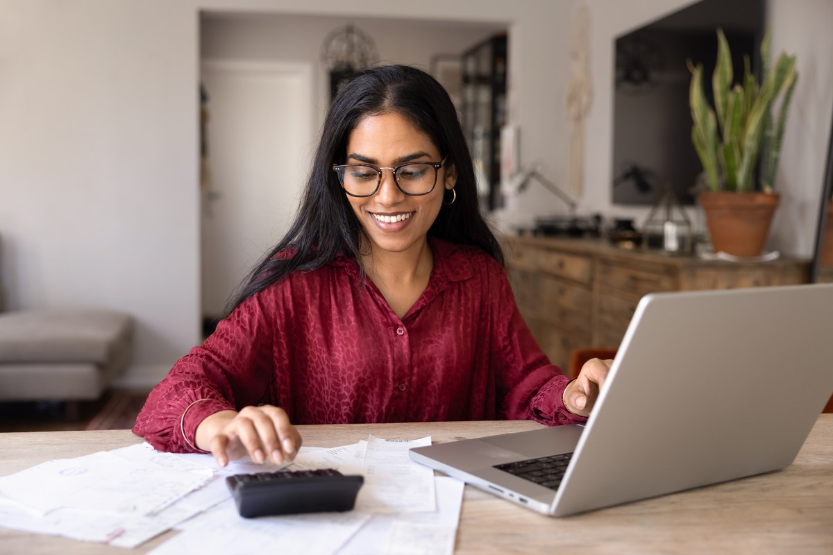 Woman with glasses using a calculator and laptop with bills at home