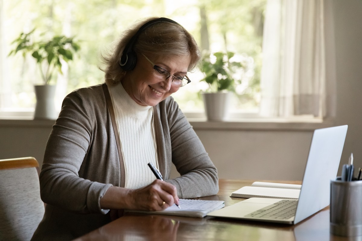 Older woman with headphones taking notes in front of a laptop at home