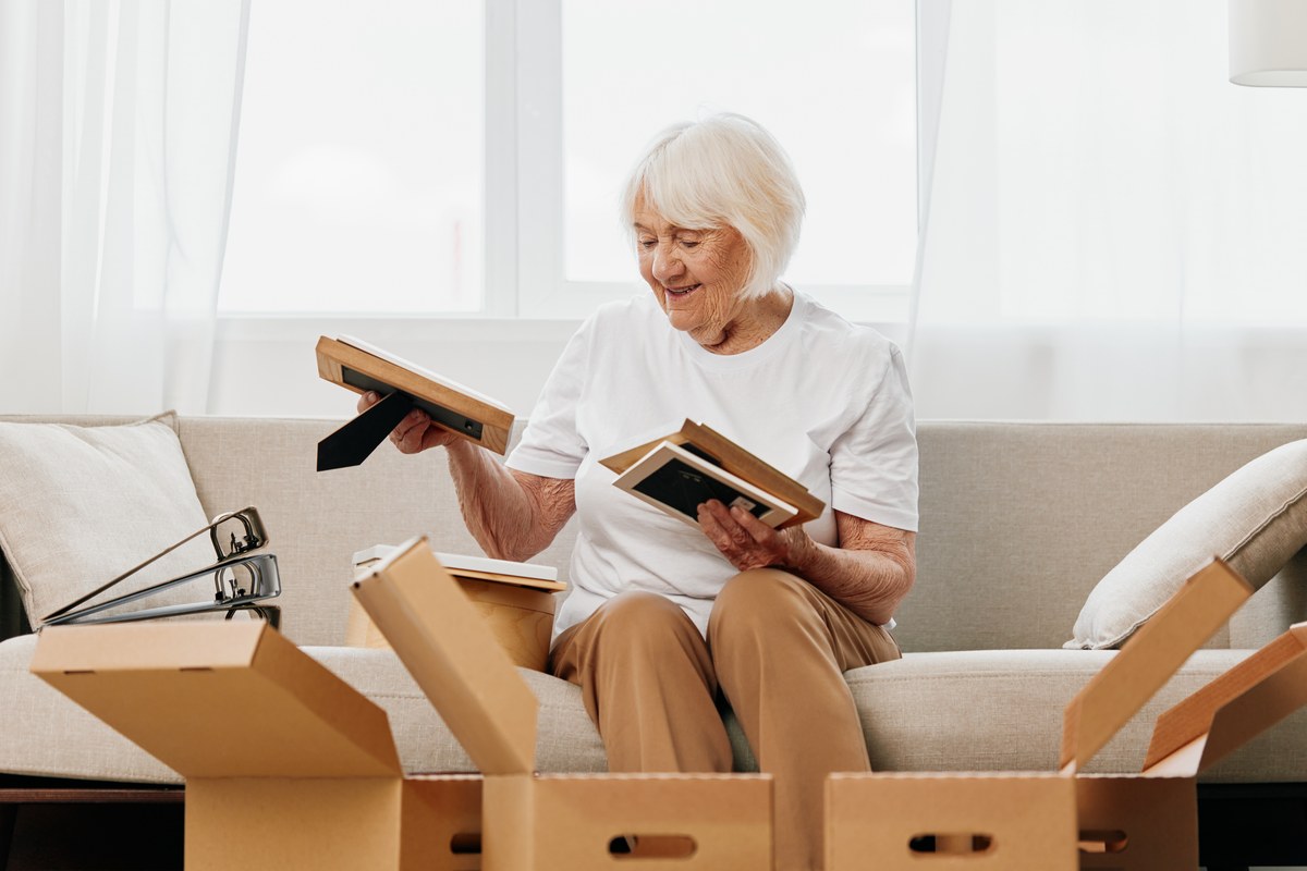 Older woman on a sofa unpacking picture frames from moving boxes