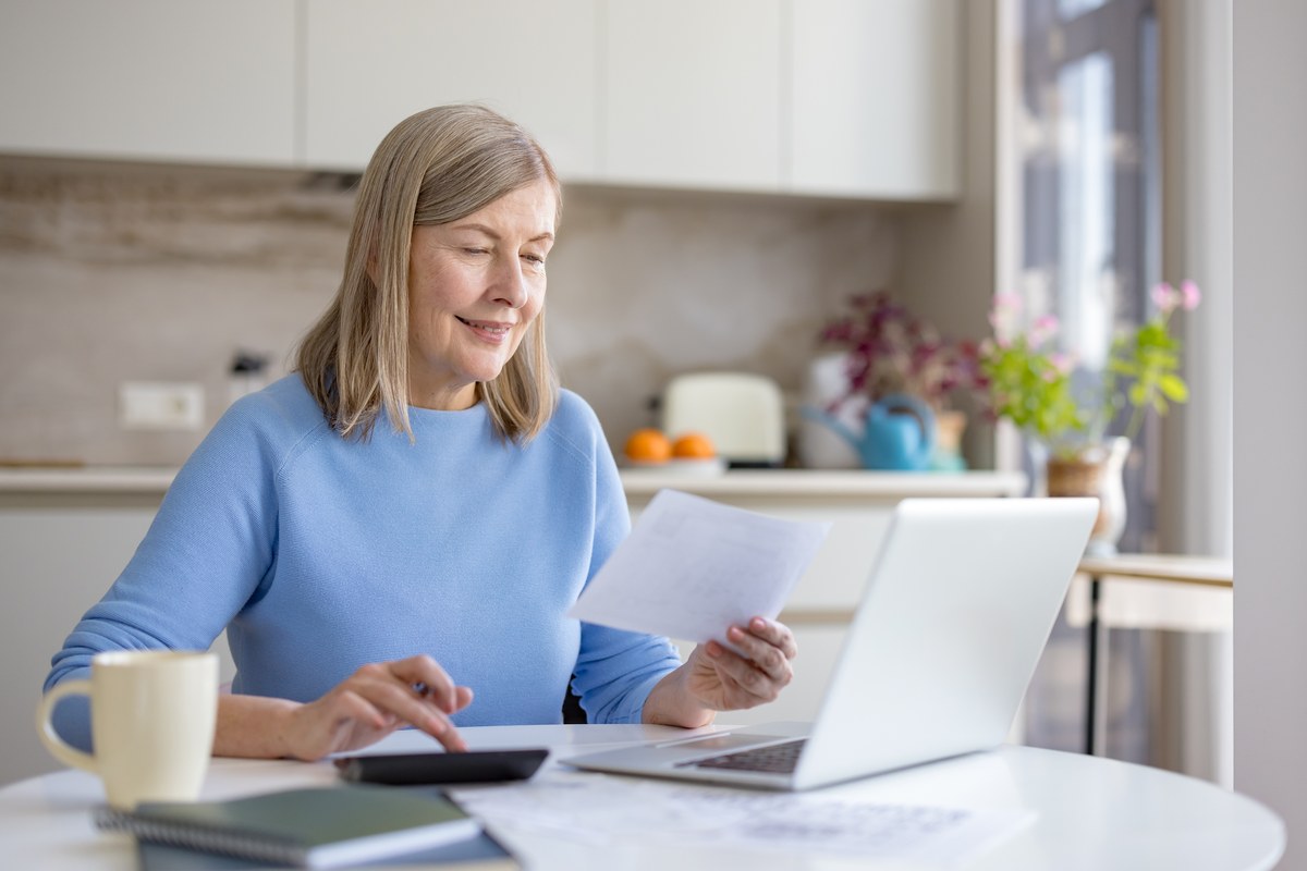 Older woman at a kitchen table reviewing papers with a laptop and calculator