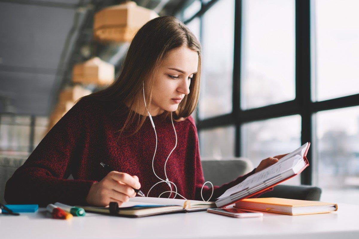 Young woman with earphones taking notes from a textbook at a desk