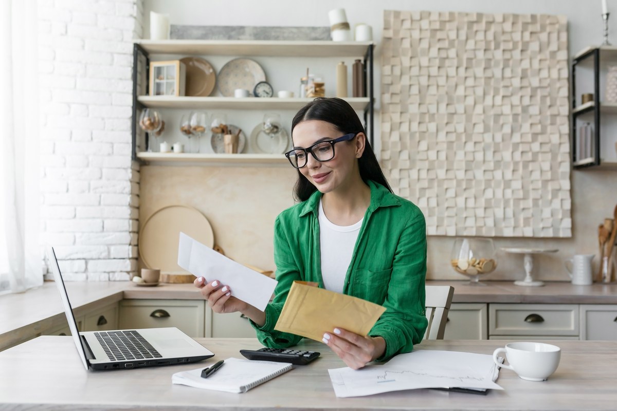Woman opening letters at a kitchen table with a laptop and calculator