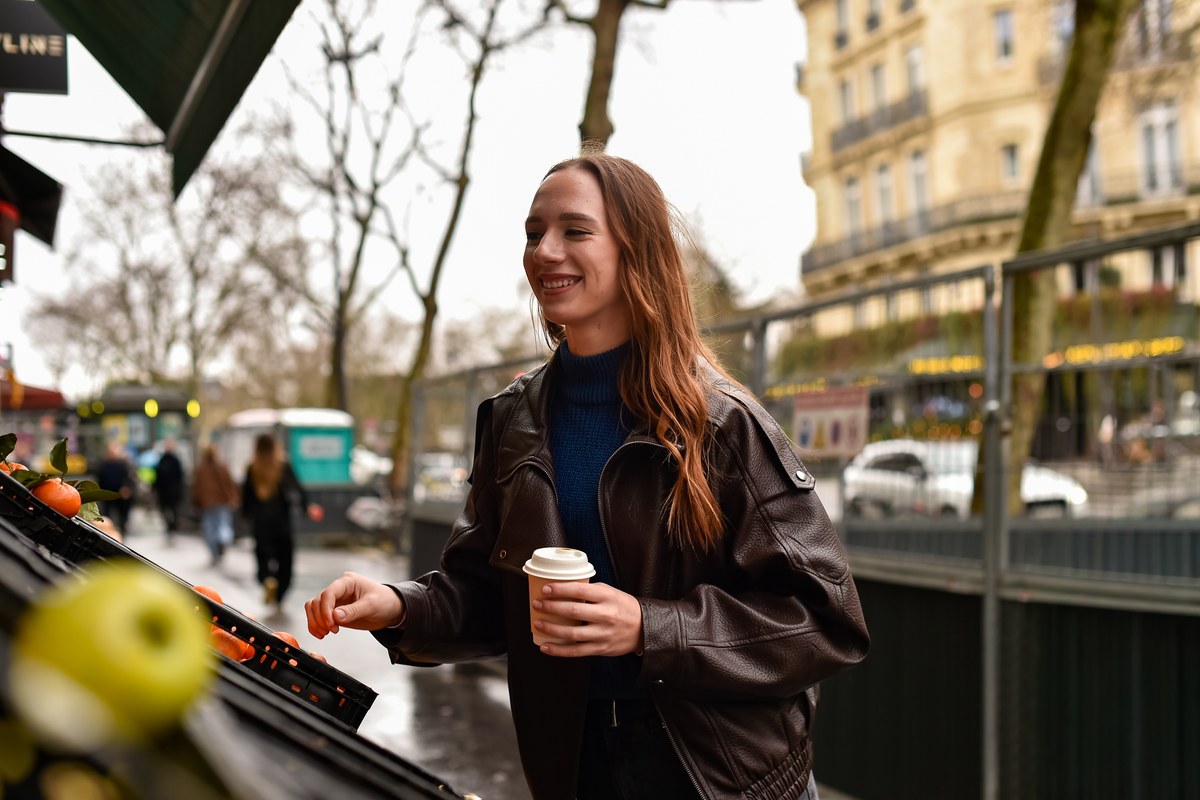 Woman with a coffee choosing fruit at an outdoor produce stall on a Paris street