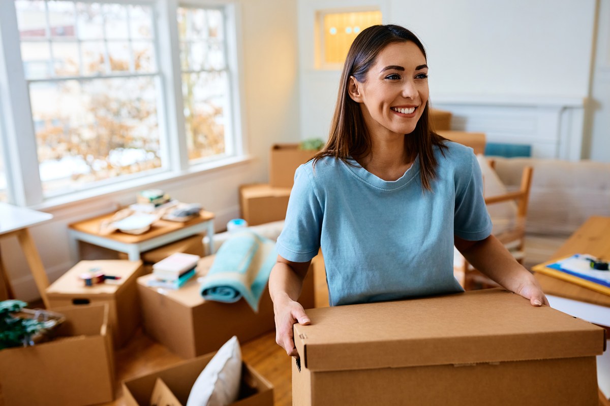 Smiling woman carrying a moving box in a room full of boxes