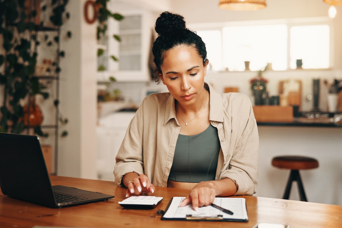 Woman at a kitchen table with a laptop, calculator and paperwork