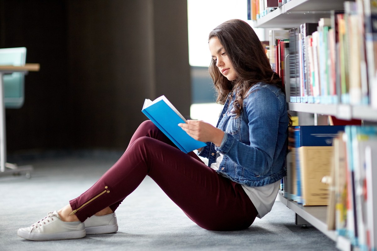 Young woman sitting on the floor of a library reading a blue book