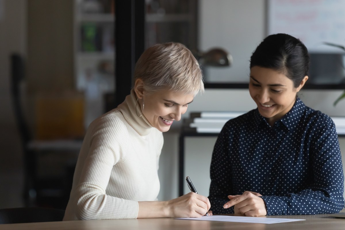 Woman signing a form at a desk while another woman helps her