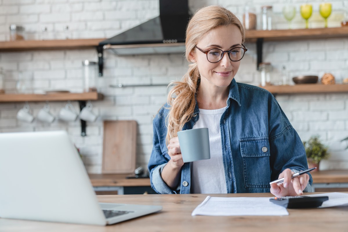 Woman with glasses and a mug going through bills with a calculator in her kitchen
