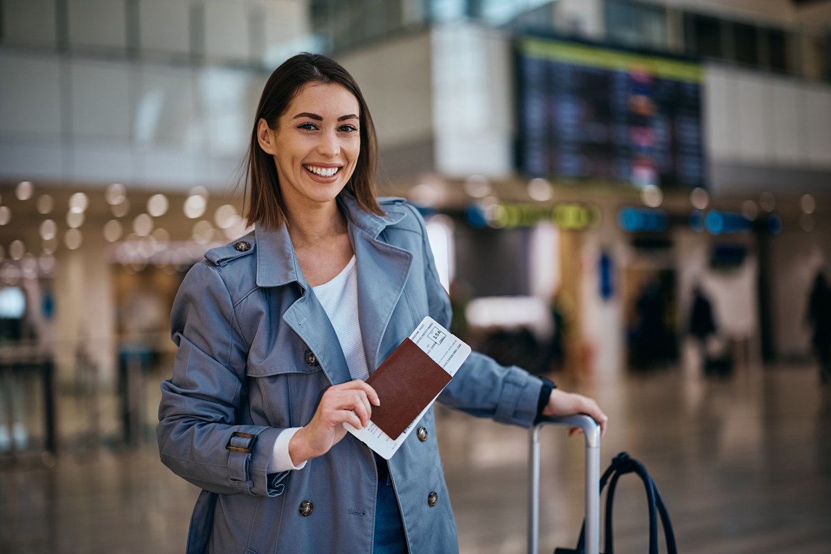 Smiling woman in a trench coat holding a passport and boarding pass at an airport