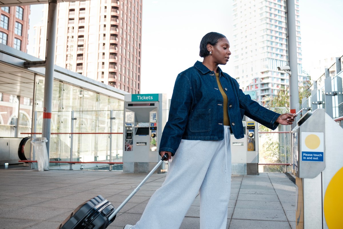 Woman with a rolling suitcase tapping her phone at a train station ticket gate