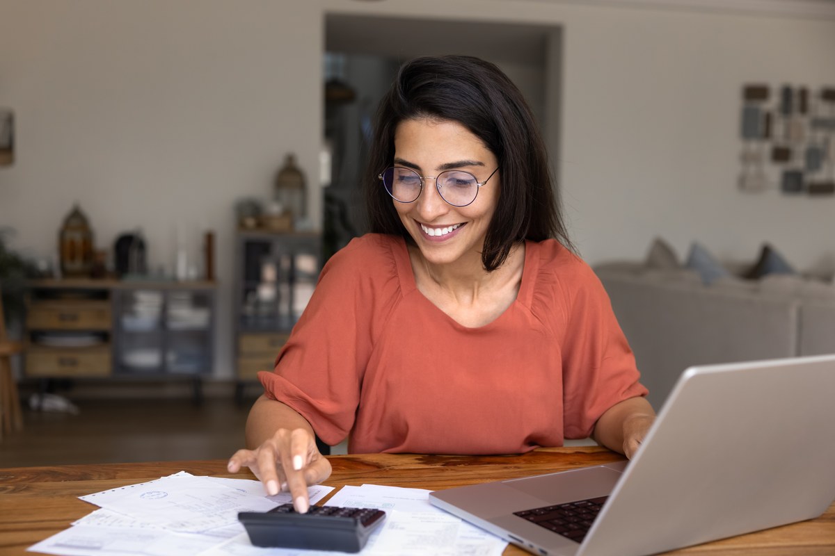Woman with glasses using a calculator and laptop with papers at home