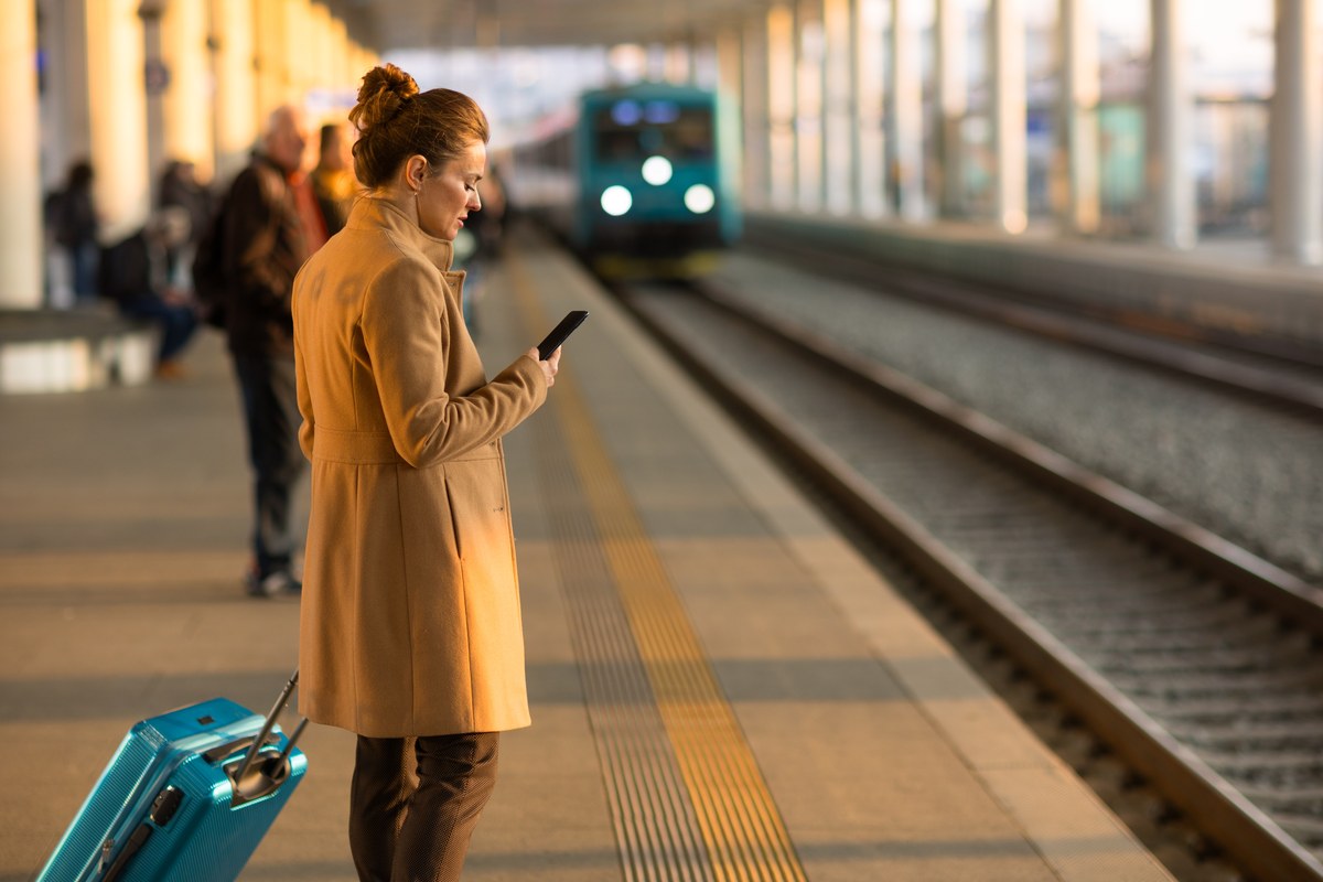 Woman with a suitcase checking her phone on a platform as a train arrives