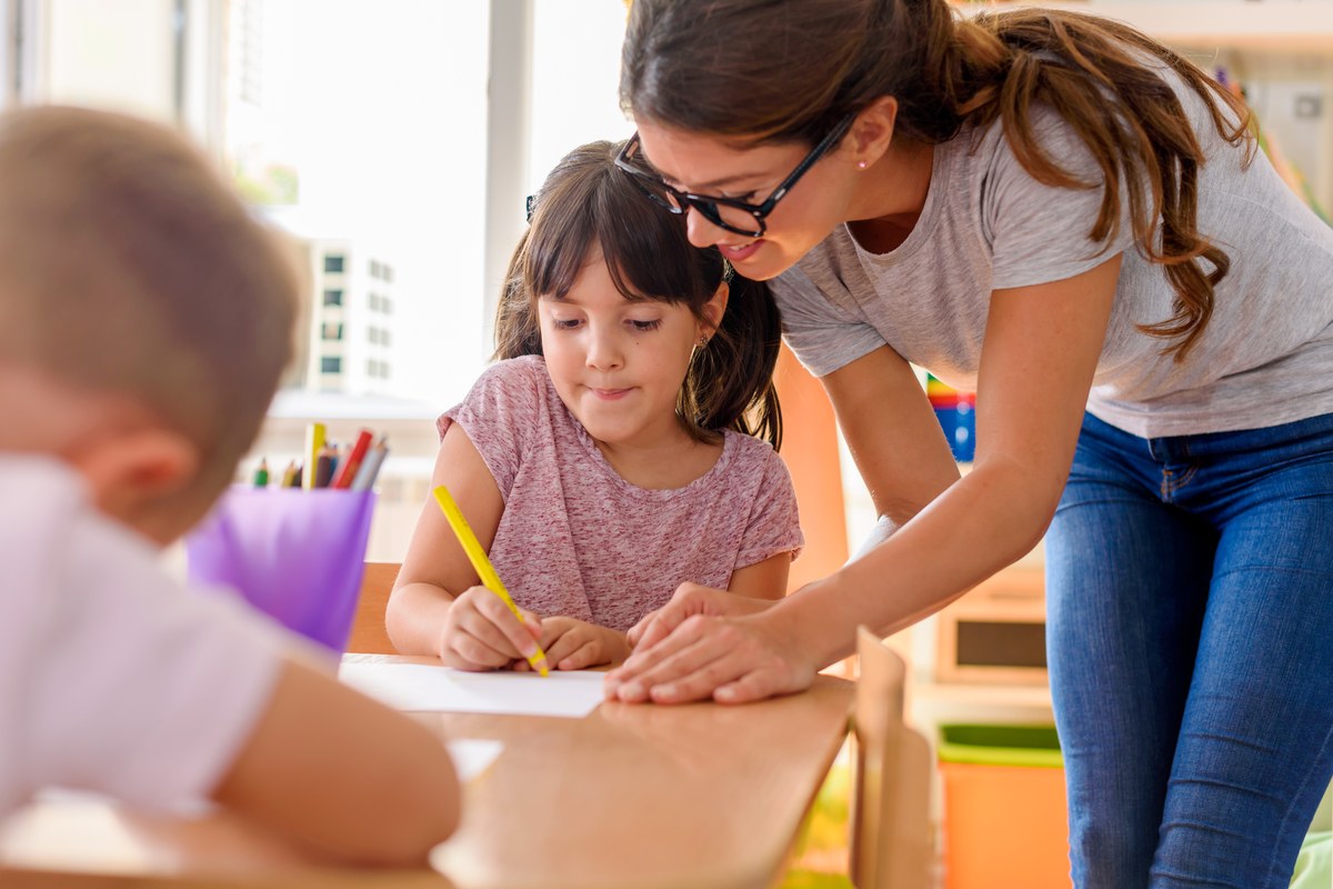 Teacher in glasses helping a girl with her drawing at a preschool table