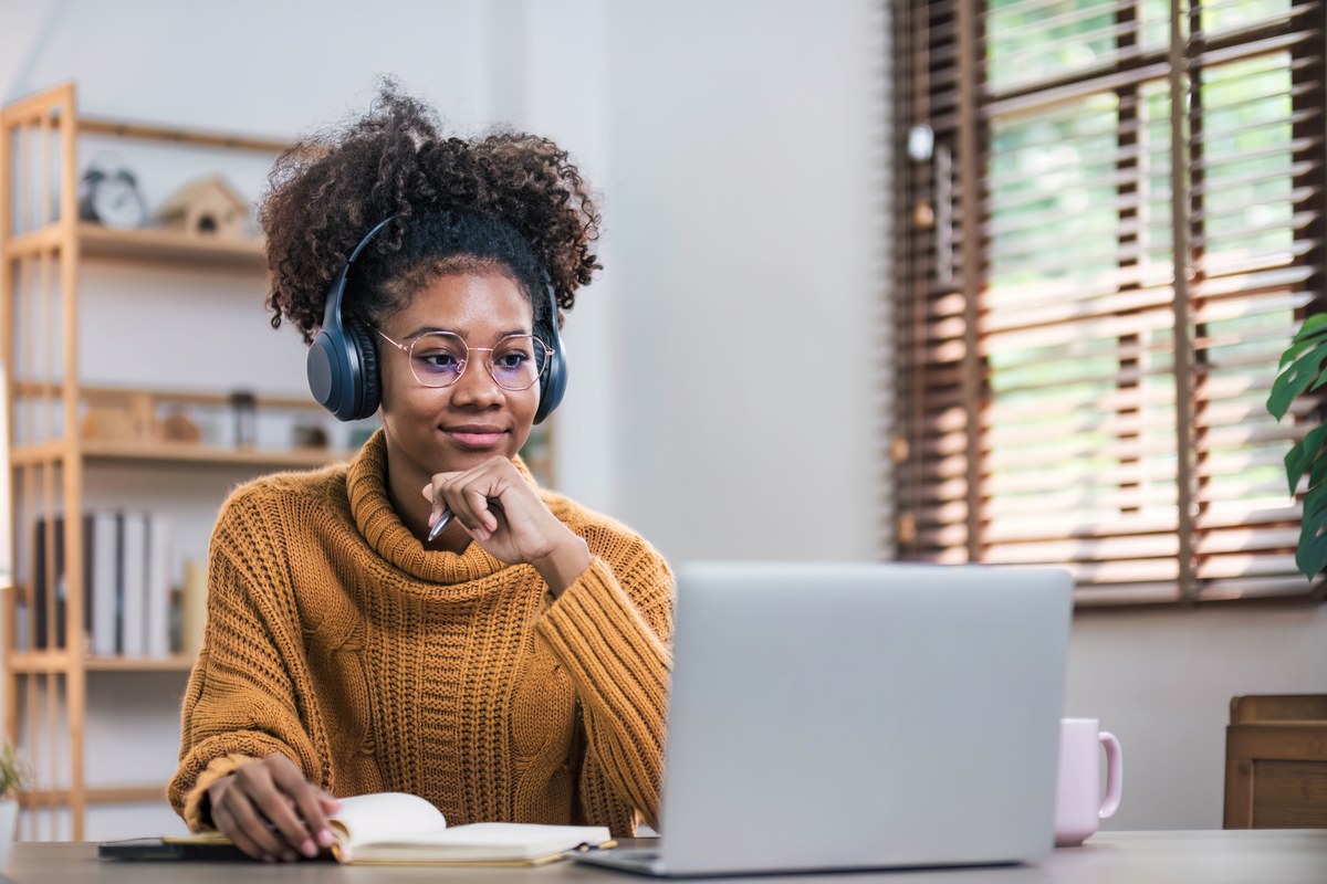Woman with headphones and glasses at a laptop with an open book at home