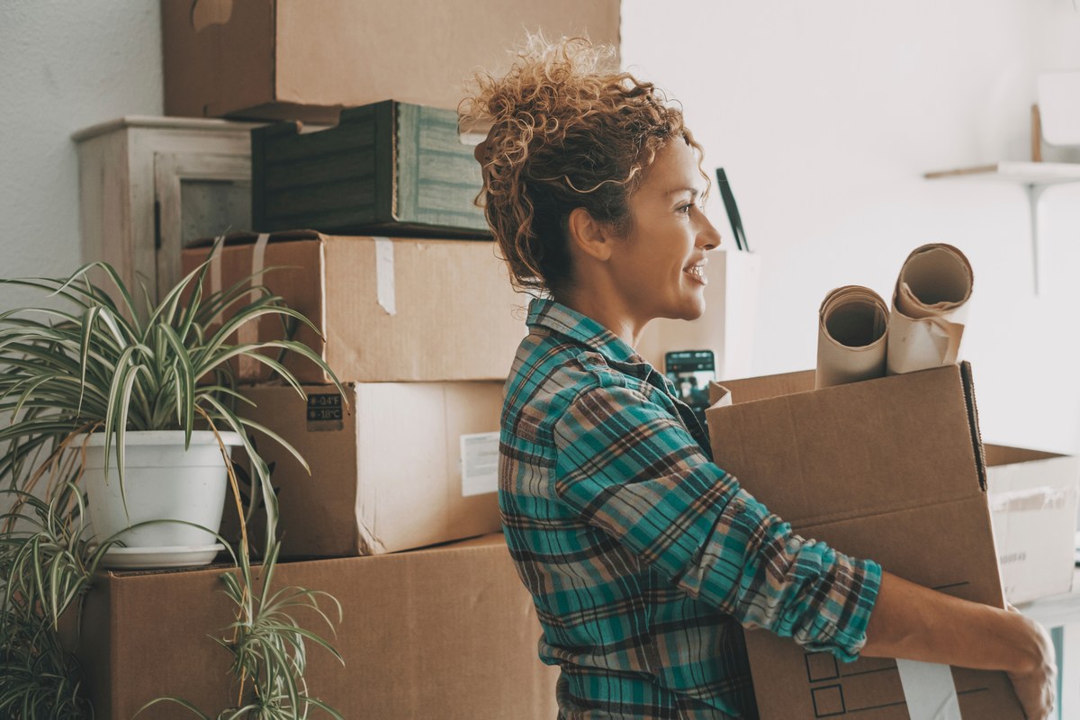 Smiling woman carrying a cardboard box among moving boxes and plants