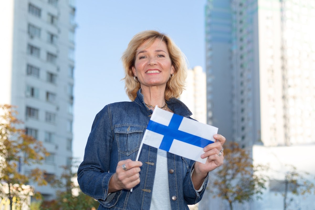 Smiling woman in a denim jacket holding a small Finnish flag in front of buildings