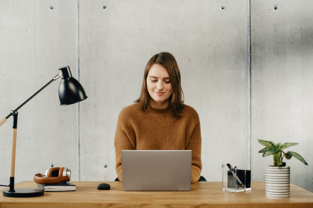 Woman working on a laptop at a wooden desk with a lamp in a concrete-walled office