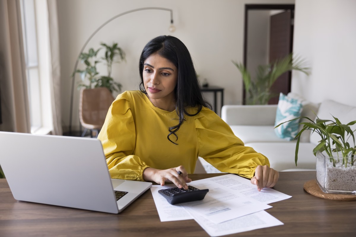 Woman in a yellow blouse using a calculator and laptop with papers at a home desk