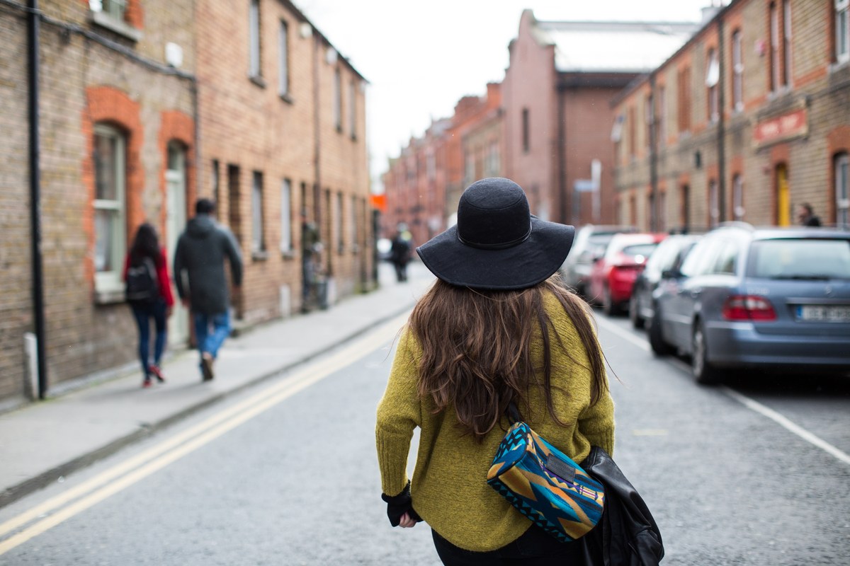 Woman in a wide-brimmed hat walking along a quiet red-brick street lined with parked cars