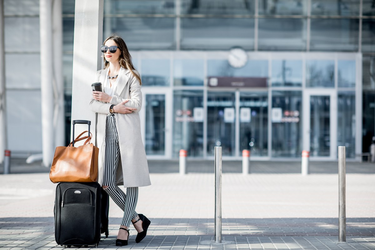 Woman with a coffee cup, handbag and rolling suitcase standing outside an airport terminal