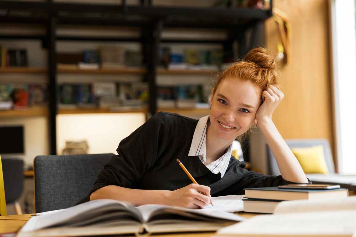Smiling red-haired woman writing notes with a book open at a library table