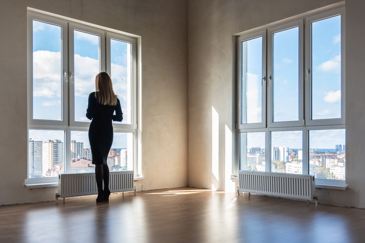 Woman standing at a tall window in an empty apartment looking out over the city