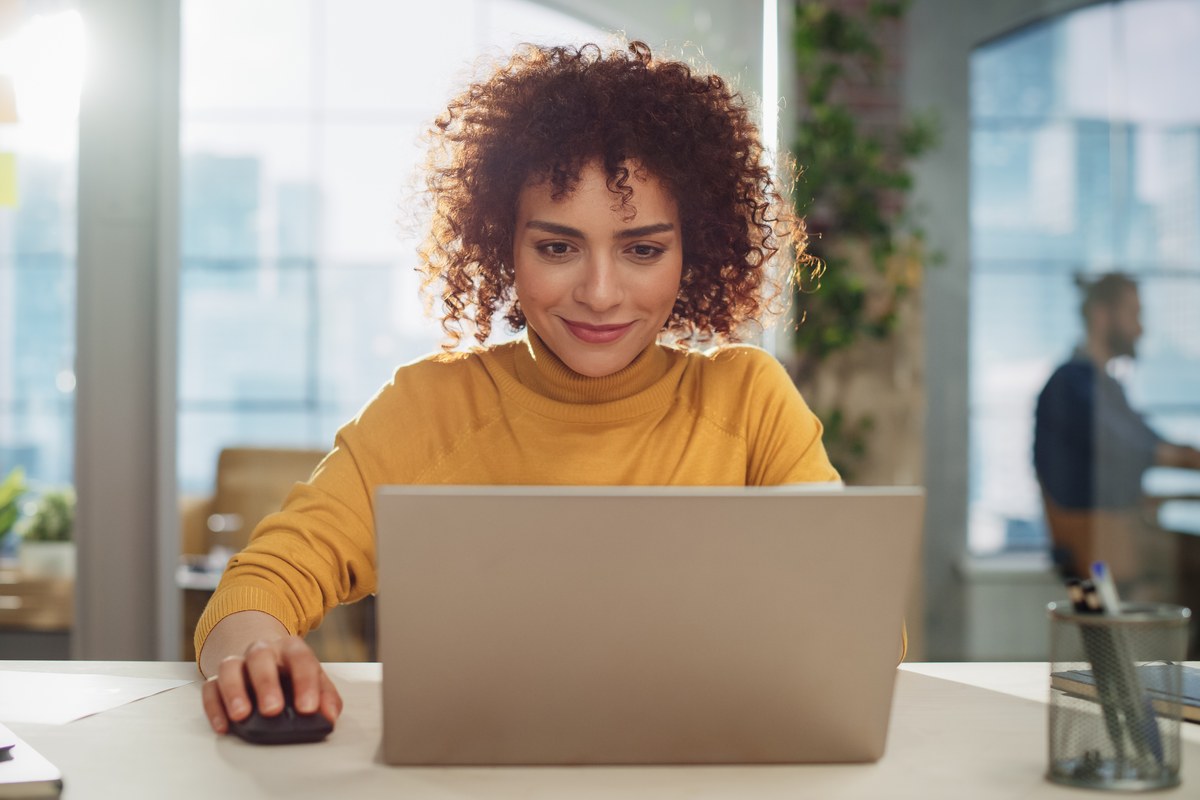 Smiling woman with curly hair working on a laptop at a desk in an open-plan office
