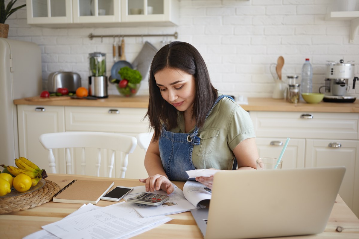 Woman using a calculator beside a laptop and papers at a kitchen table