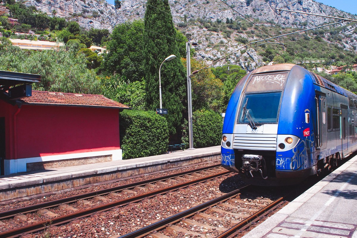 Blue regional train at a small station below wooded hills at Eze