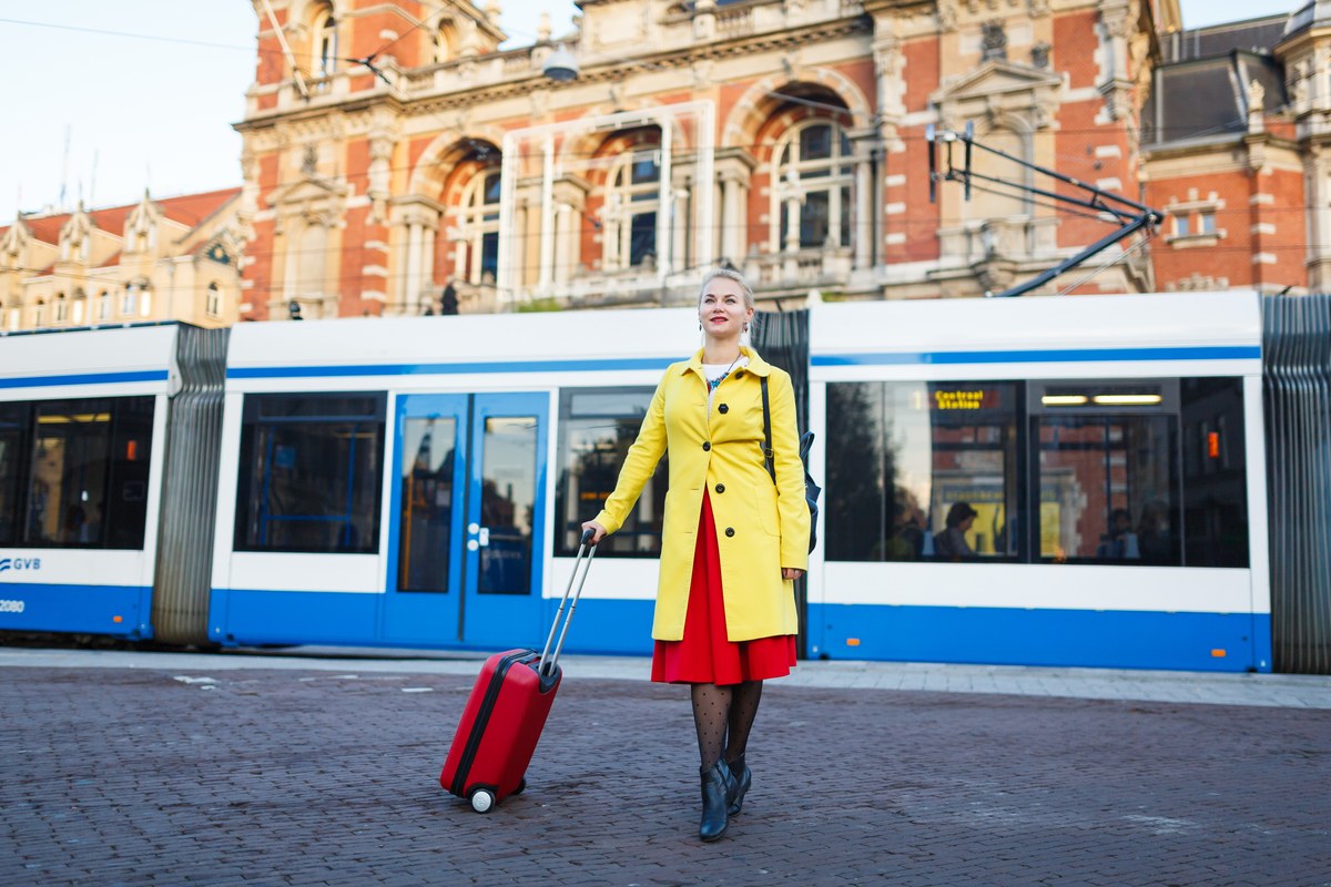 Woman in a yellow coat pulling a red suitcase in front of a tram in Amsterdam