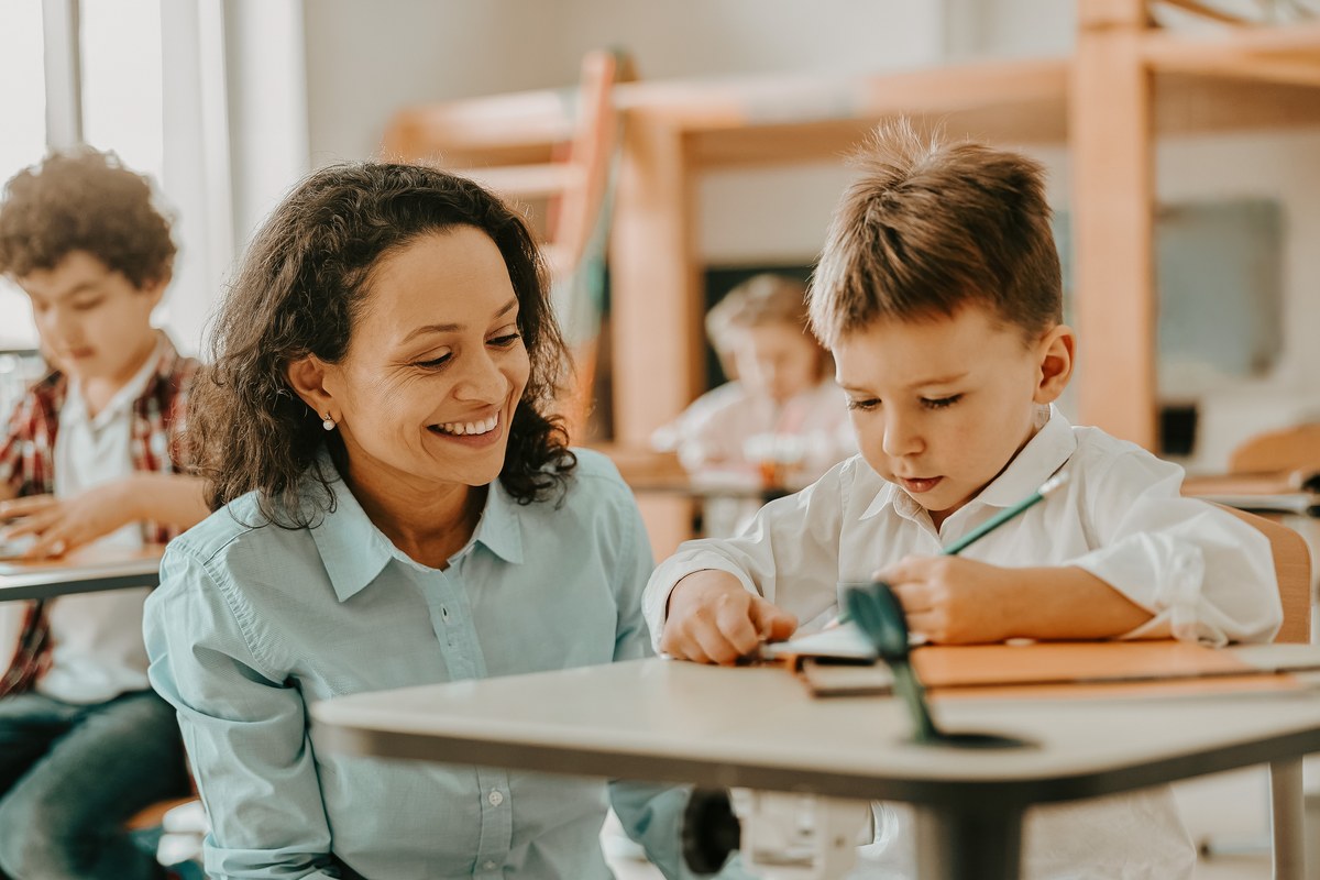 Teacher smiling beside a boy writing at his desk in a classroom