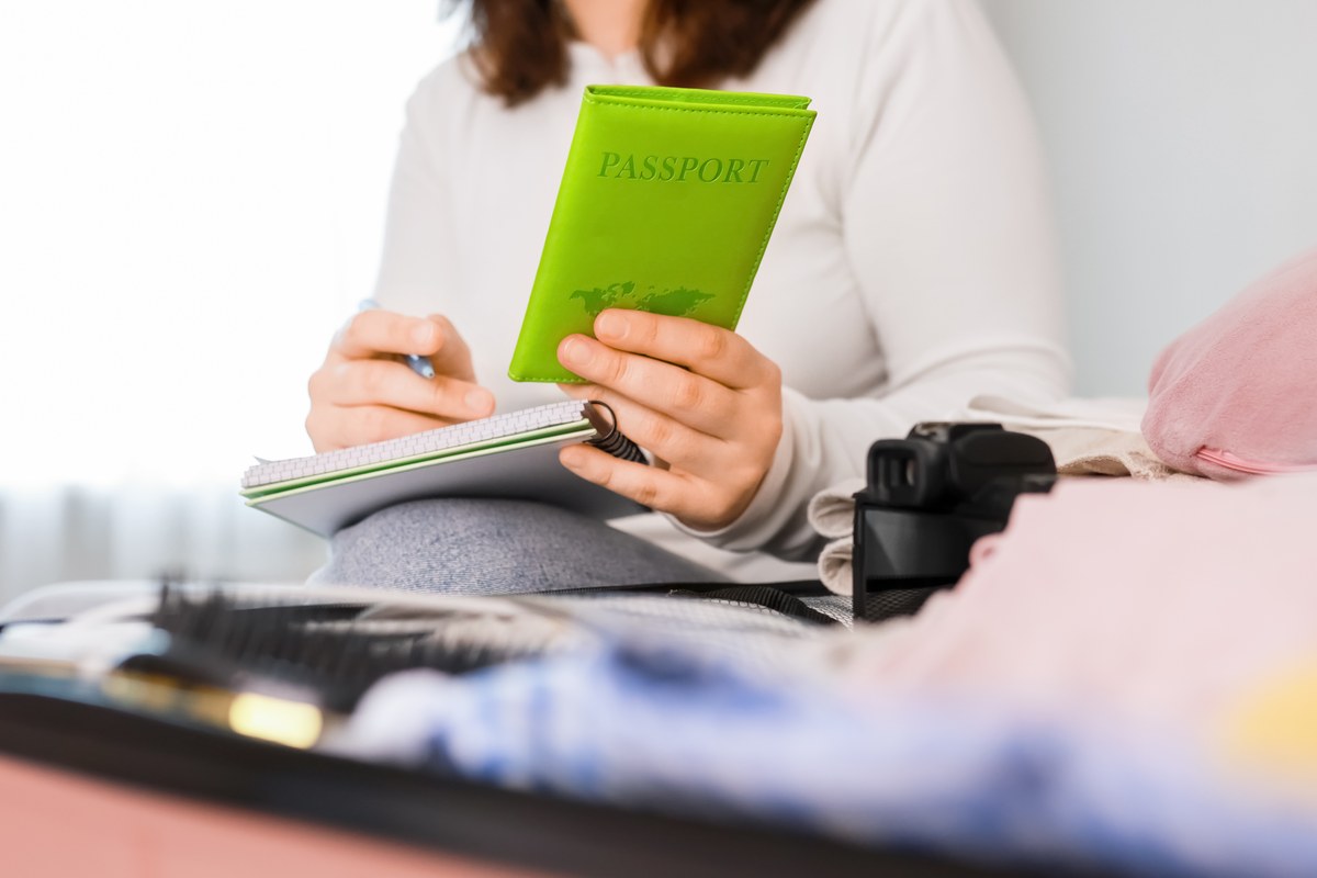 Woman writing a packing list while holding a green passport beside a suitcase