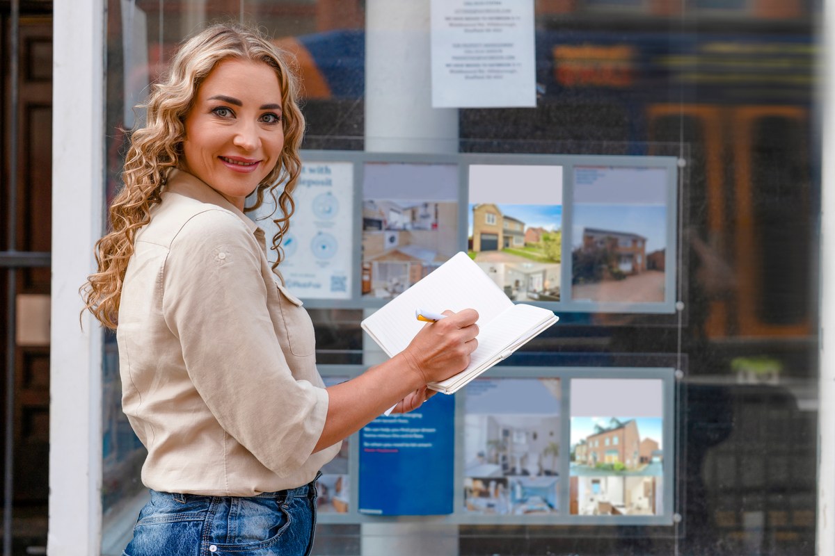 Woman writing in a notebook while looking at property listings in an estate agent window
