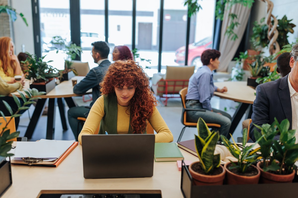Woman with curly red hair working on a laptop in a plant-filled open office