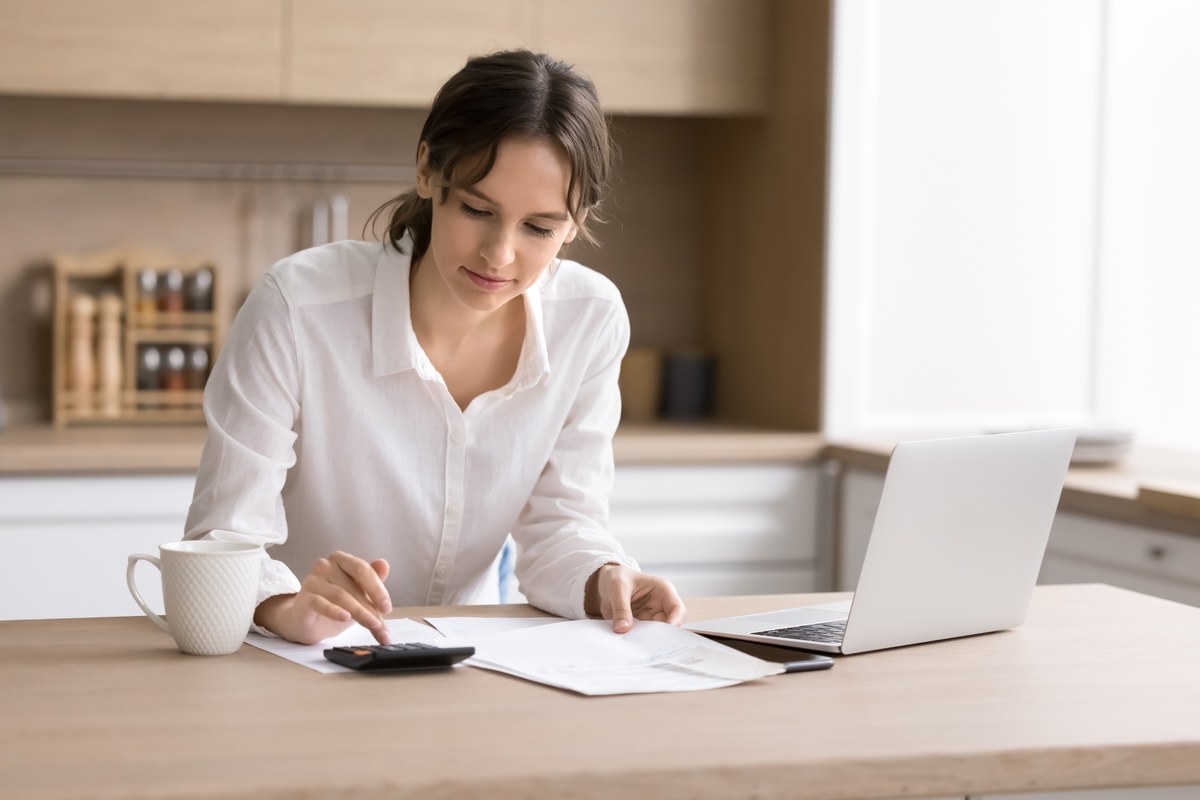 Woman using a calculator with papers and a laptop at a kitchen table