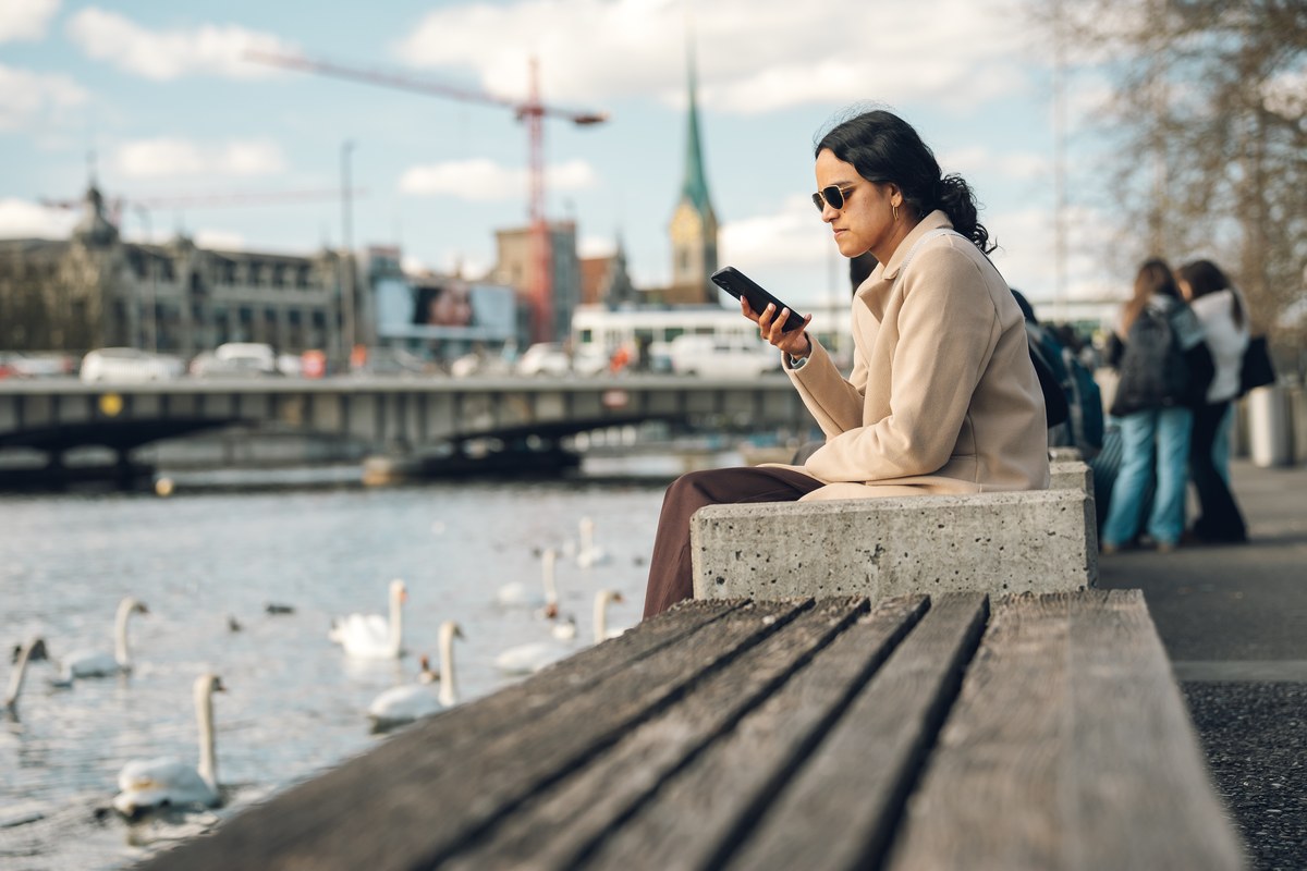 A woman in a beige coat checks her phone on a bench by the Limmat river in Zurich