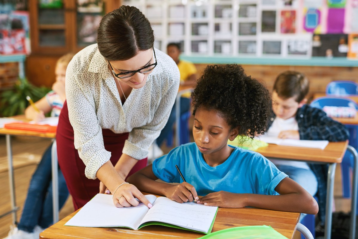 A teacher leans over a desk to help a girl writing in an exercise book in a classroom