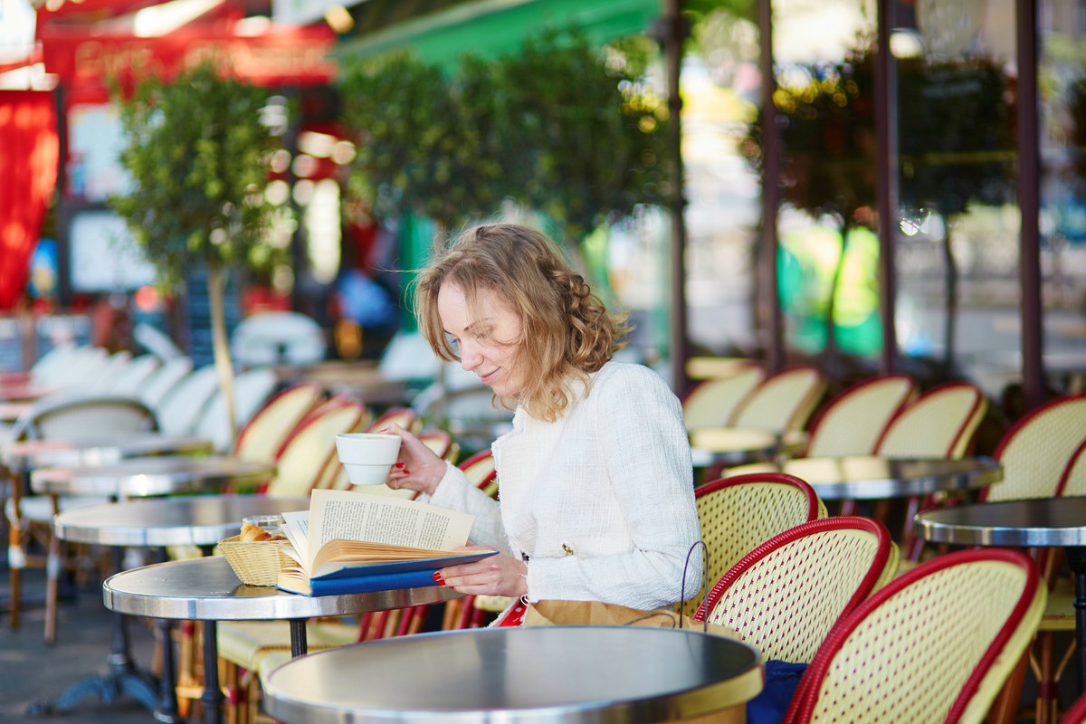 A woman reads a book with a cup of coffee at a Paris cafe terrace with wicker chairs