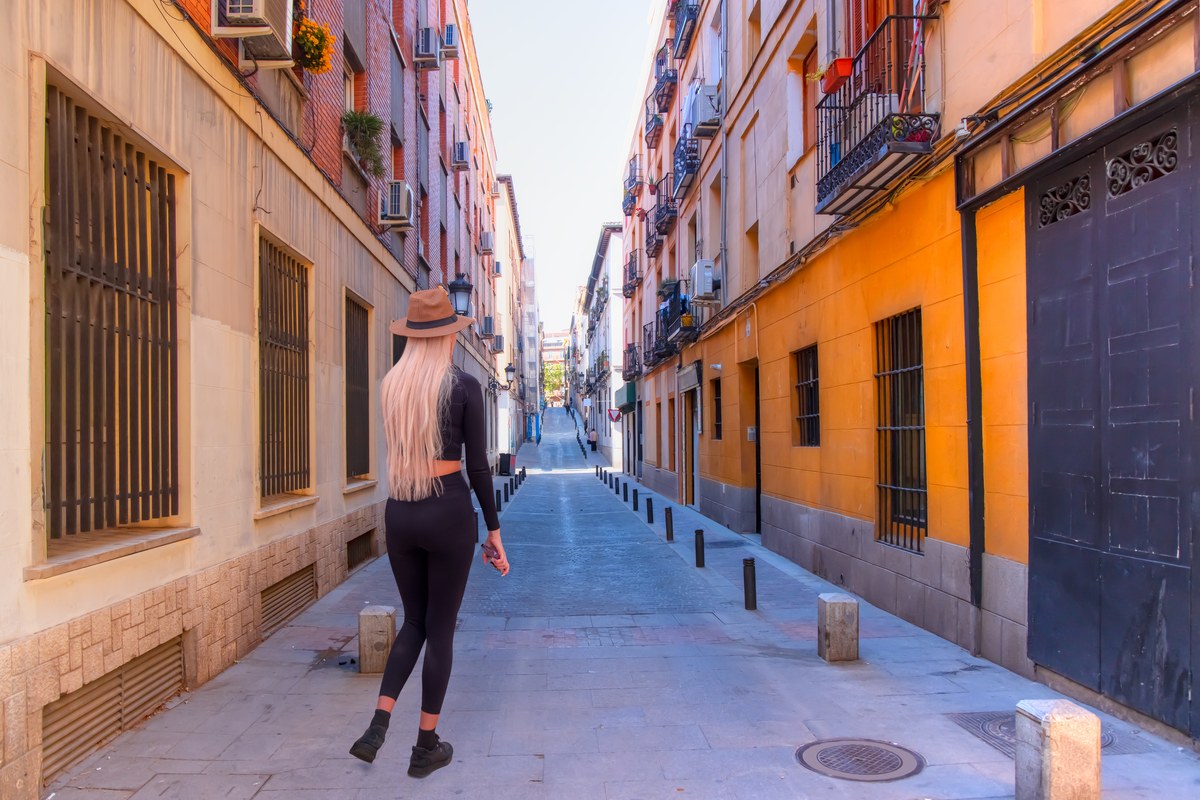 A woman in a hat walks down a narrow street of balconied apartment buildings in Madrid