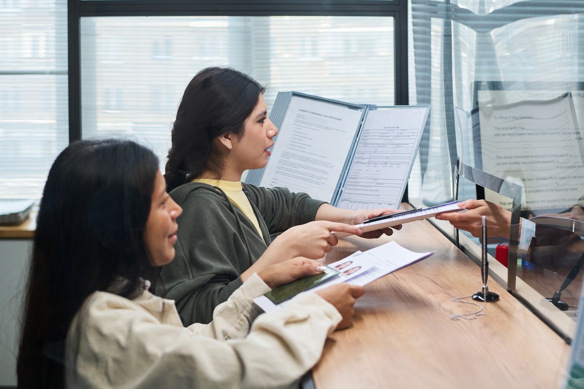 Two women hand documents to a clerk through a glass partition at an office counter