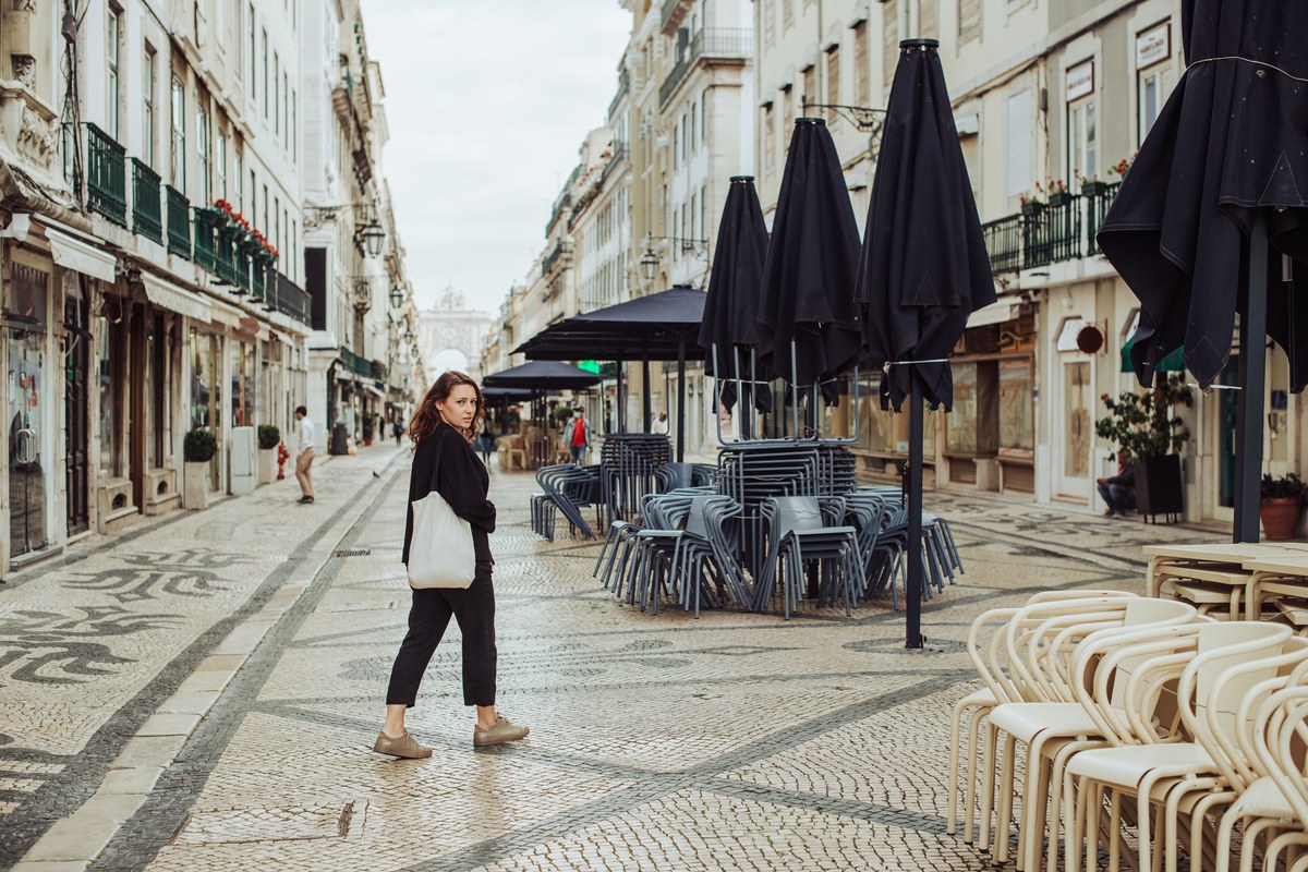Woman with a tote bag walking past a closed café terrace on a cobbled Lisbon street