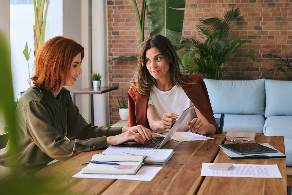 Two women working with a laptop and papers at a desk in a green office